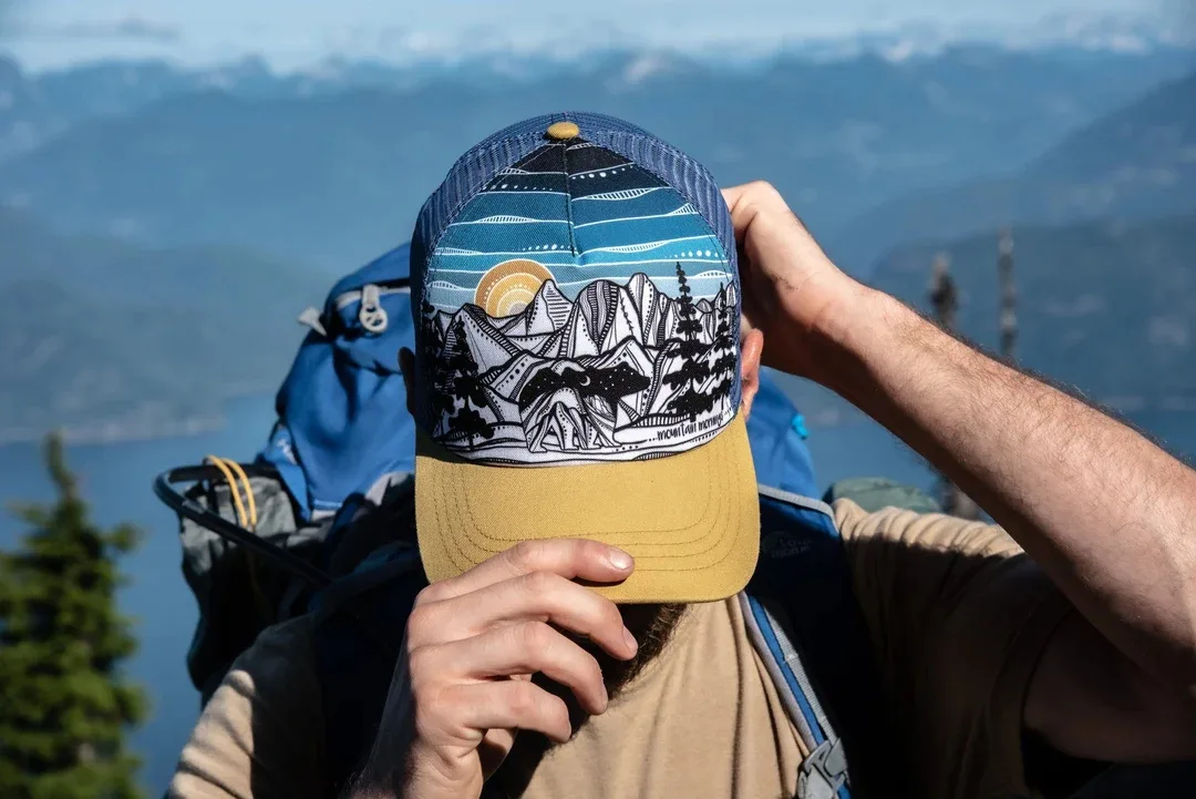 Close up of the front of an Ambler Mountain Works baseball cap with a scene of mountains on it. It is worn by a man who has been hiking in the mountains.