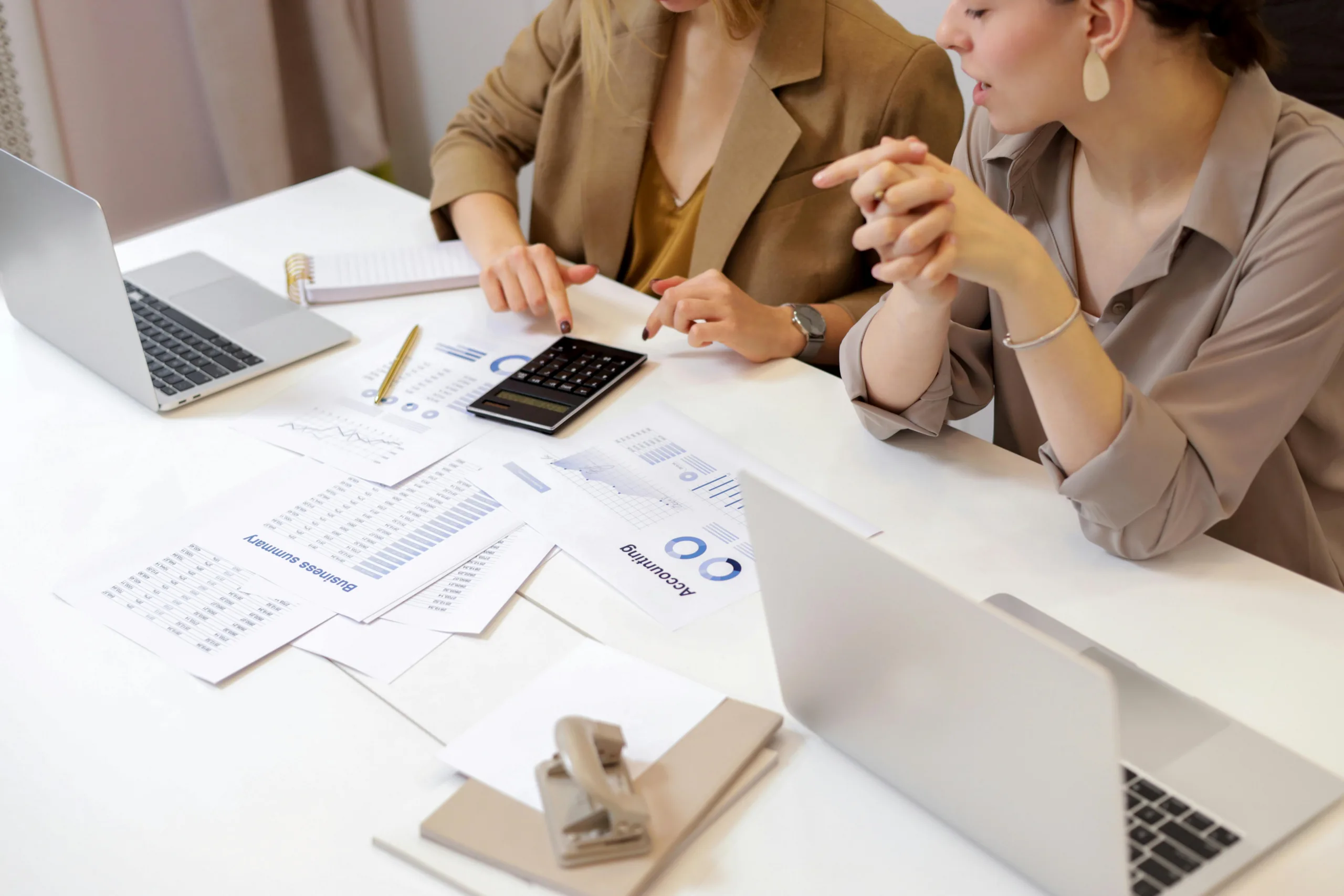 Two business people work with laptops and a calculator with accounting documents spread out in front of them.