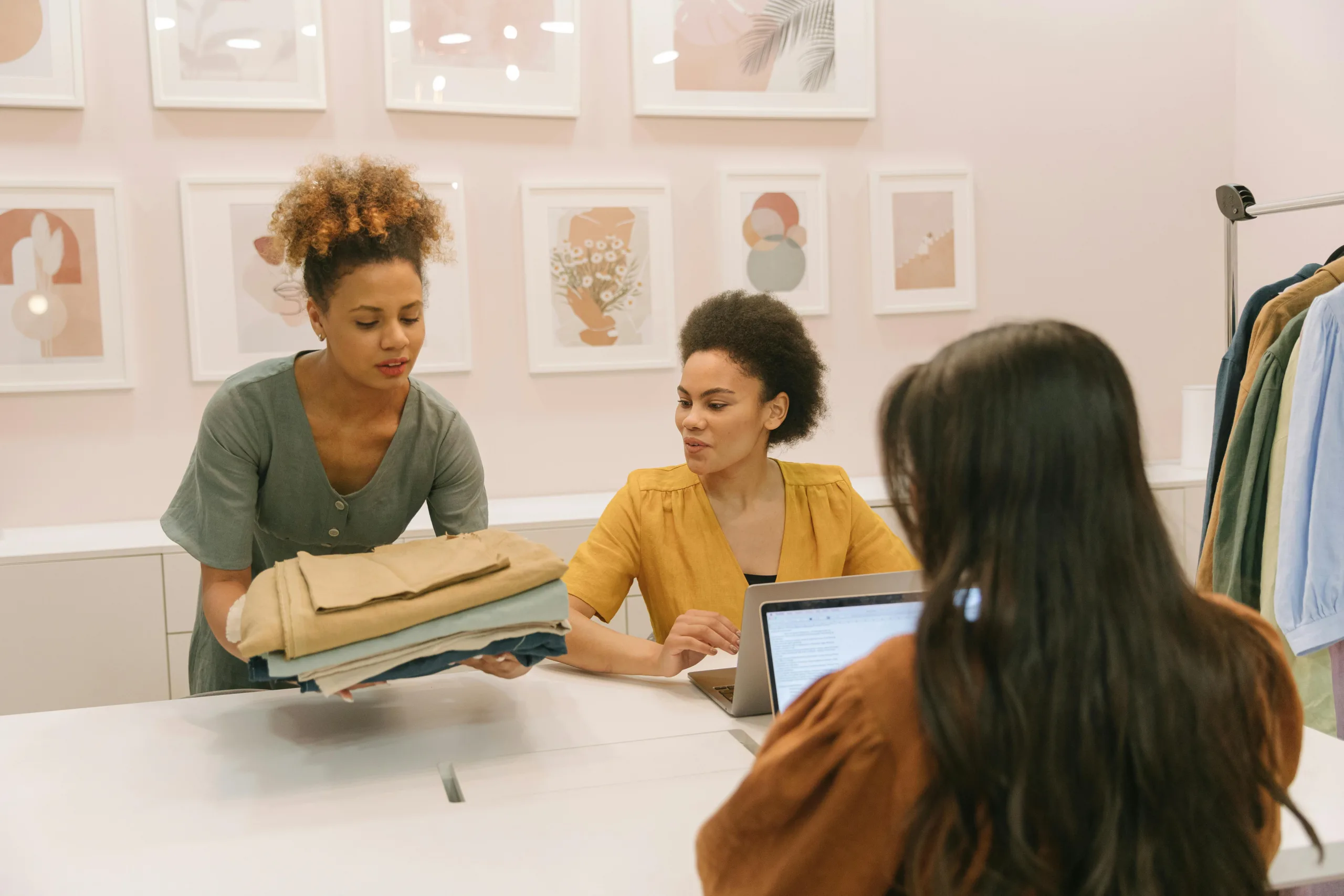 Three women are sitting at a table. Two of them are working on laptops and the third is carrying several fabric samples and placing them on the table. In the background is a rack of clothing.