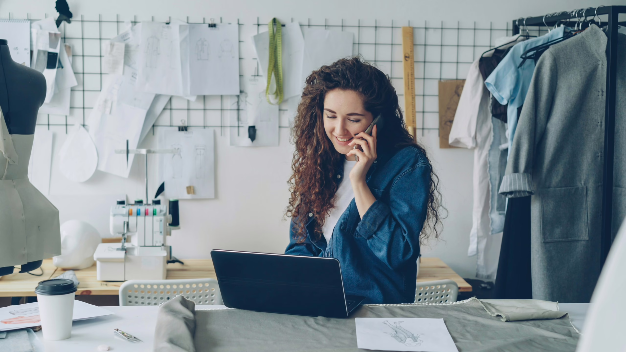 A young woman talks on the phone while looking at a laptop; around her are clothes and sewing supplies, including a sewing machine.
